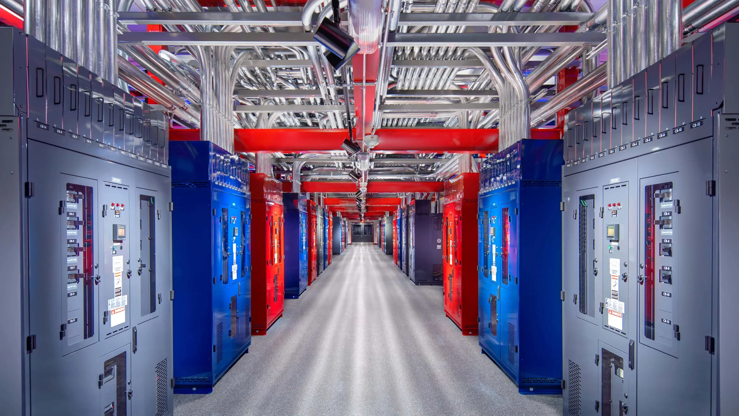 n data center electrical room: a long corridor flanked by rows of red and blue power cabinets, overhead cable trays and conduit piping, clean floor and bright lighting highlighting industrial infrastructure.