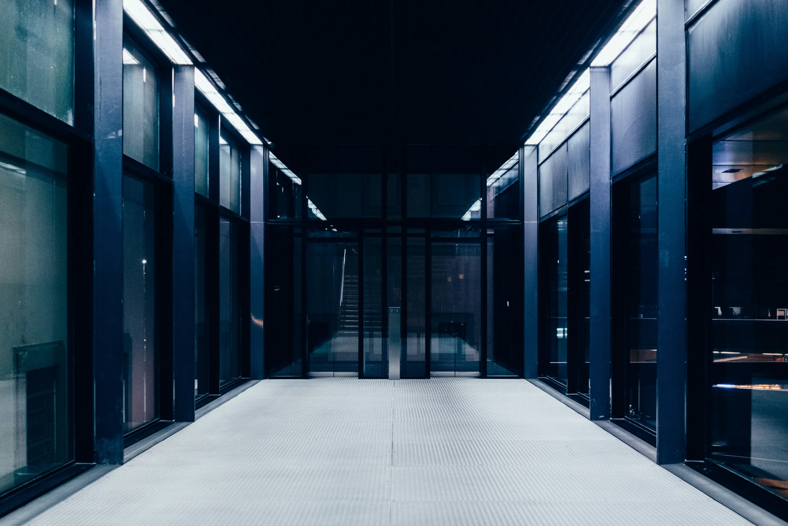 Modern, empty corridor with glass walls and bright overhead lighting, leading to a glass door with a staircase visible in the background.