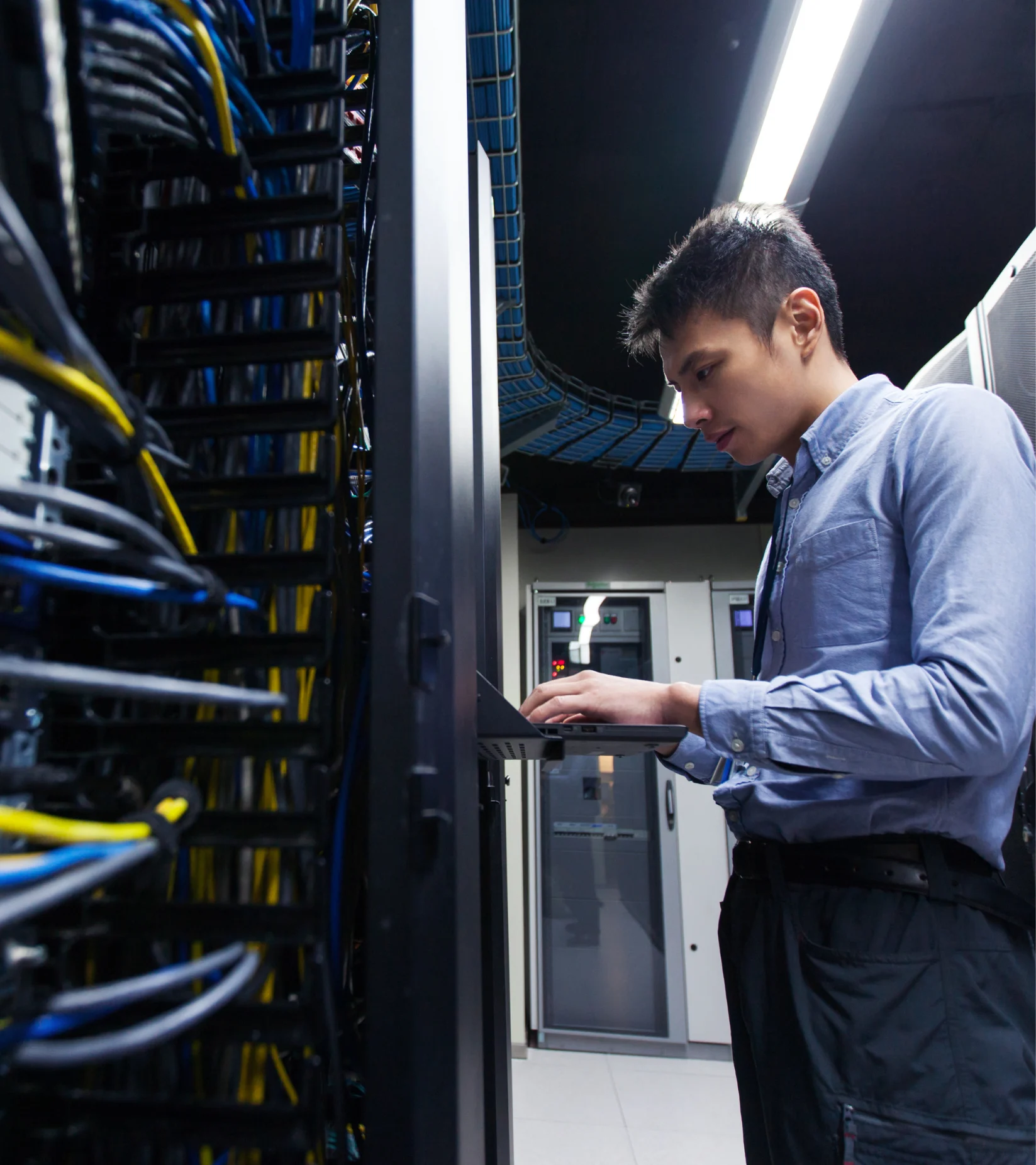 IT technician working on a laptop in a data center, surrounded by server racks and network cables.